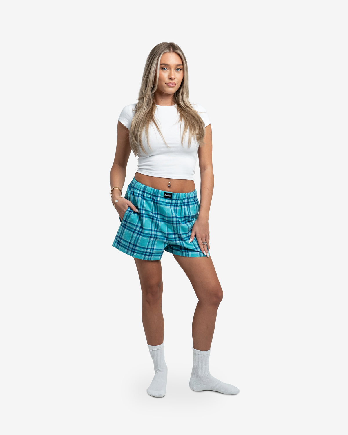 Frau trägt Hot Pants in der Farbe Blueberry. Die Shorts haben ein lockeres, hüftbetontes Design und ein auffälliges Karomuster in Hellblau und Dunkelblau. Der elastische Bund mit leichtem Faltenwurf sorgt für eine bequeme Passform. Vorne mittig ist das schwarze Schlafi-Label aufgenäht. Kombiniert mit weißem Crop-Top und weißen Socken.