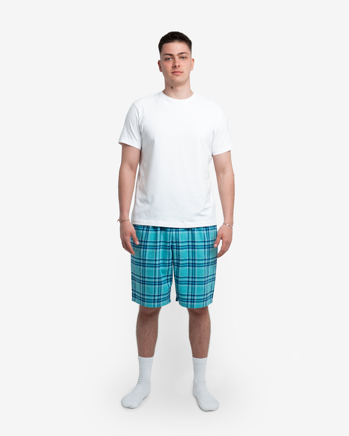 Ein junger Mann steht vor hellem Hintergrund und trägt ein weißes T-Shirt sowie eine kurz geschnittene Schlafhose in der Farbe „Blueberry“ mit türkis-blauem Karomuster. Die Shorts wirken lässig, locker geschnitten und enden knapp über dem Knie.