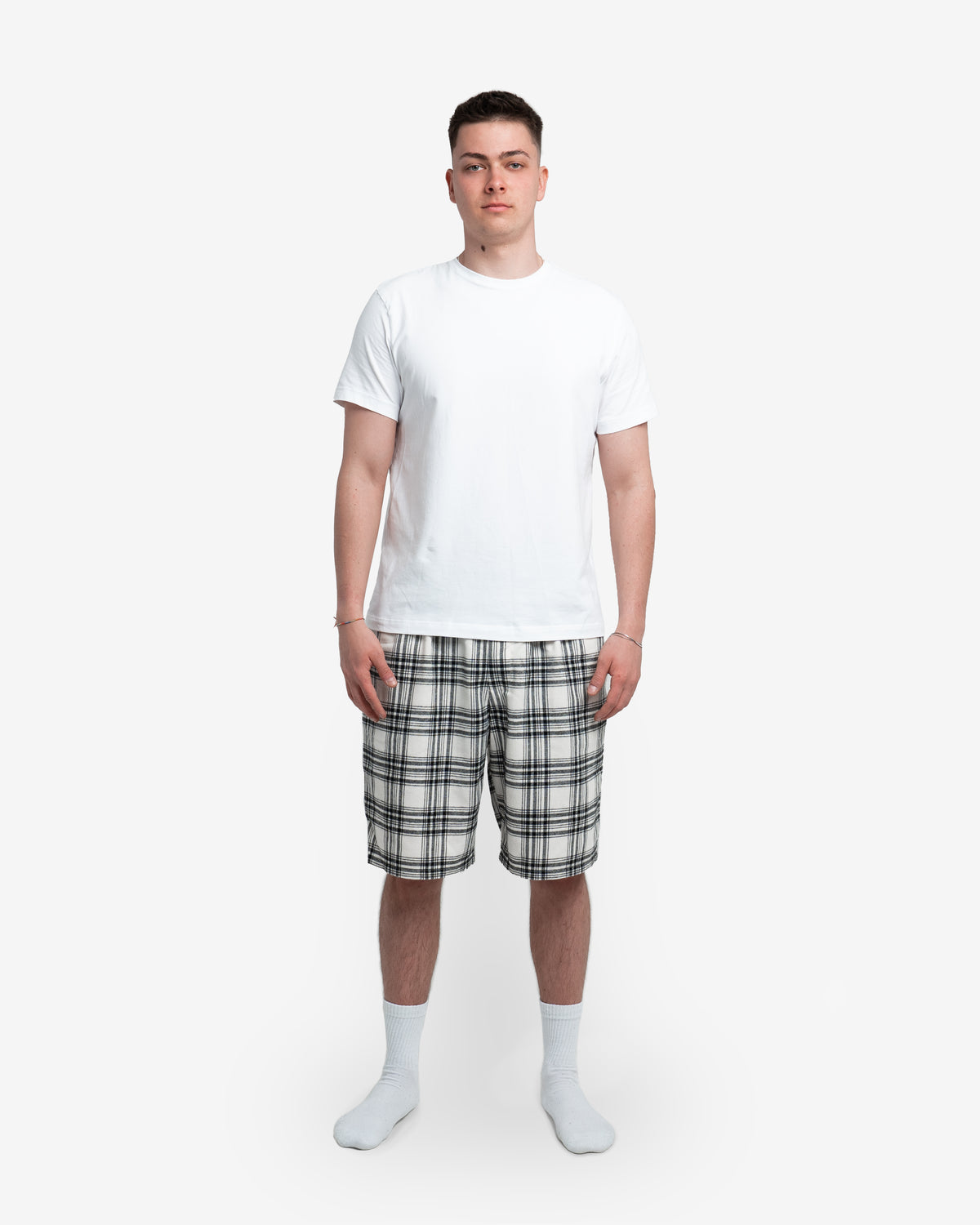 Männliches Model steht frontal zur Kamera und trägt die cremefarbene Schorti mit schwarzem Karomuster. Kombiniert mit weißem T-Shirt und weißen Socken. Hände locker an den Seiten. Hintergrund neutral weiß.