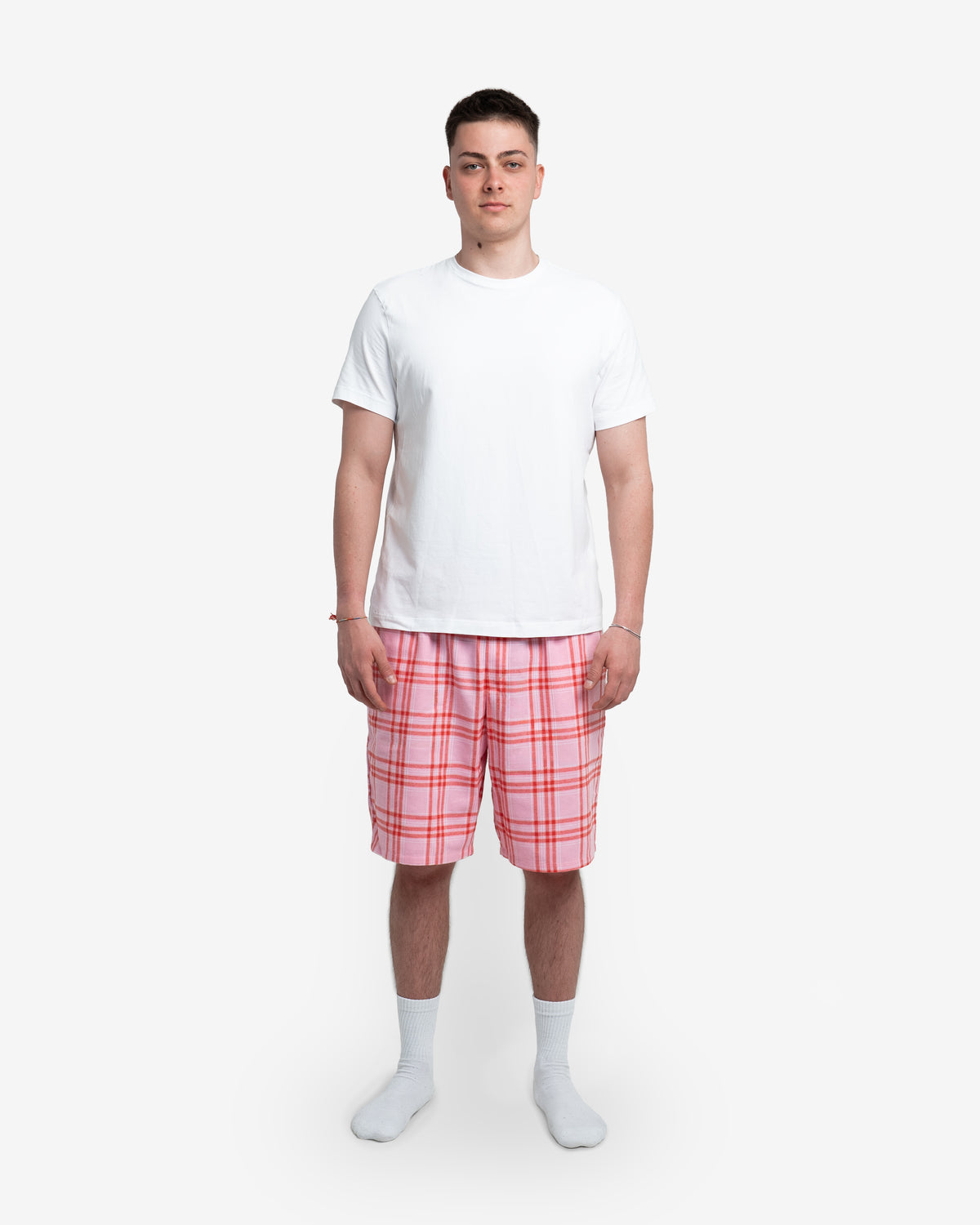 Männliches Model steht frontal zur Kamera und trägt die Schorti in Dragonfruit – ein kräftiger Pinkton mit schwarzem Karomuster. Kombiniert mit weißem T-Shirt und weißen Socken. Arme locker seitlich, neutraler weißer Hintergrund.