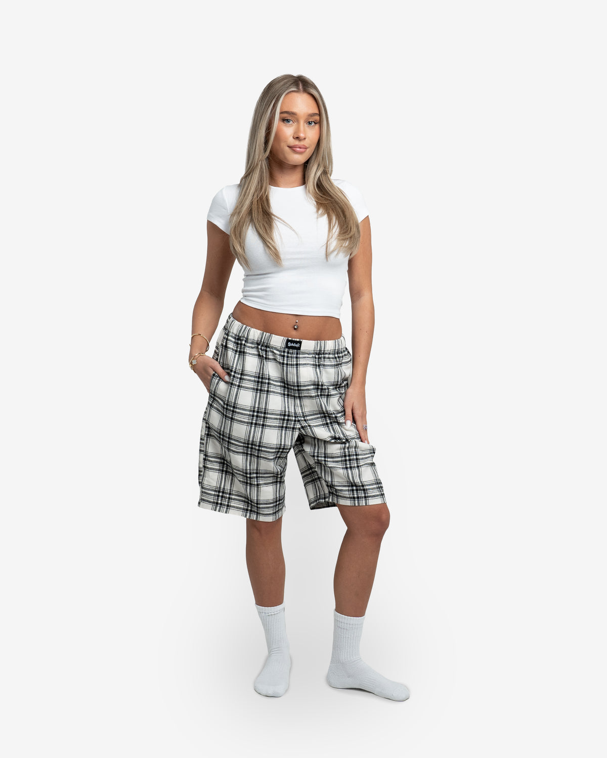 Weibliches Model steht frontal zur Kamera und trägt die Schorti in Creme mit schwarzem Karomuster. Kombiniert mit kurzem weißen Top und weißen Socken. Eine Hand in der Tasche, die andere am Oberschenkel. Neutraler weißer Hintergrund.