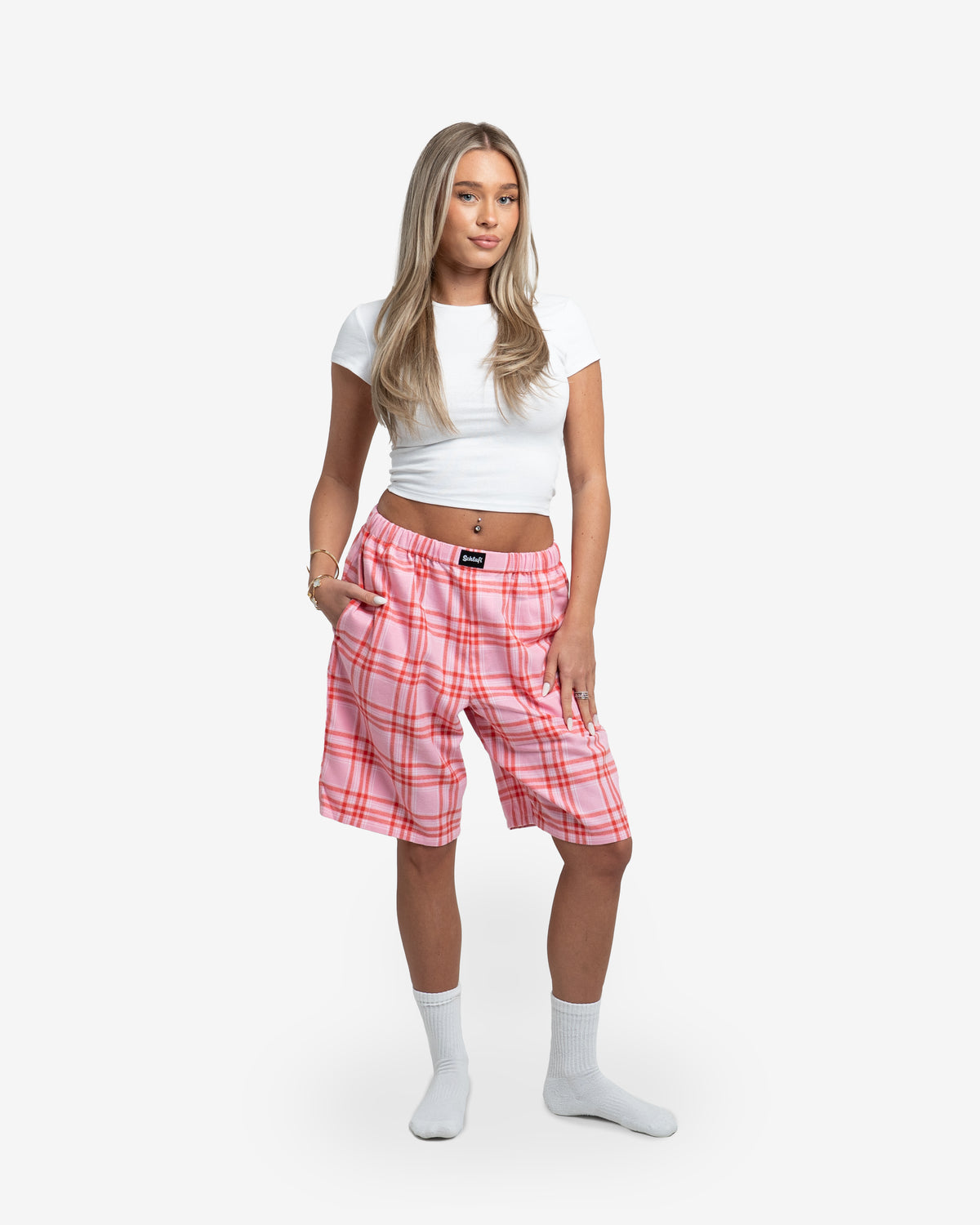 Weibliches Model steht frontal zur Kamera und trägt die Schorti in Dragonfruit mit schwarzem Karomuster. Kombiniert mit weißem, bauchfreiem T-Shirt und weißen Socken. Eine Hand locker in der Tasche, andere am Oberschenkel. Hintergrund weiß.