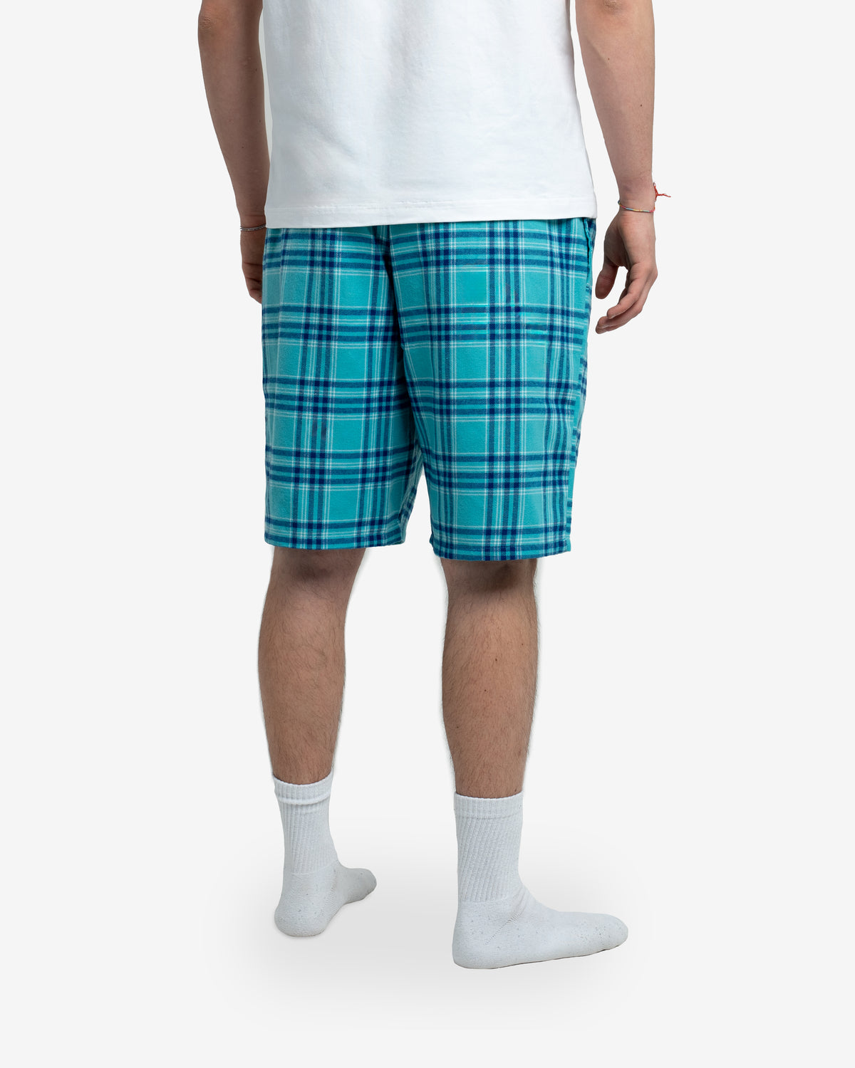 Modell steht mit dem Rücken zur Kamera und zeigt die Rückseite der Schorti in Blueberry mit blauem Karomuster. Weißes T-Shirt darüber, weiße Socken an den Füßen. Fokus auf Schnitt und Rückansicht der Shorts.