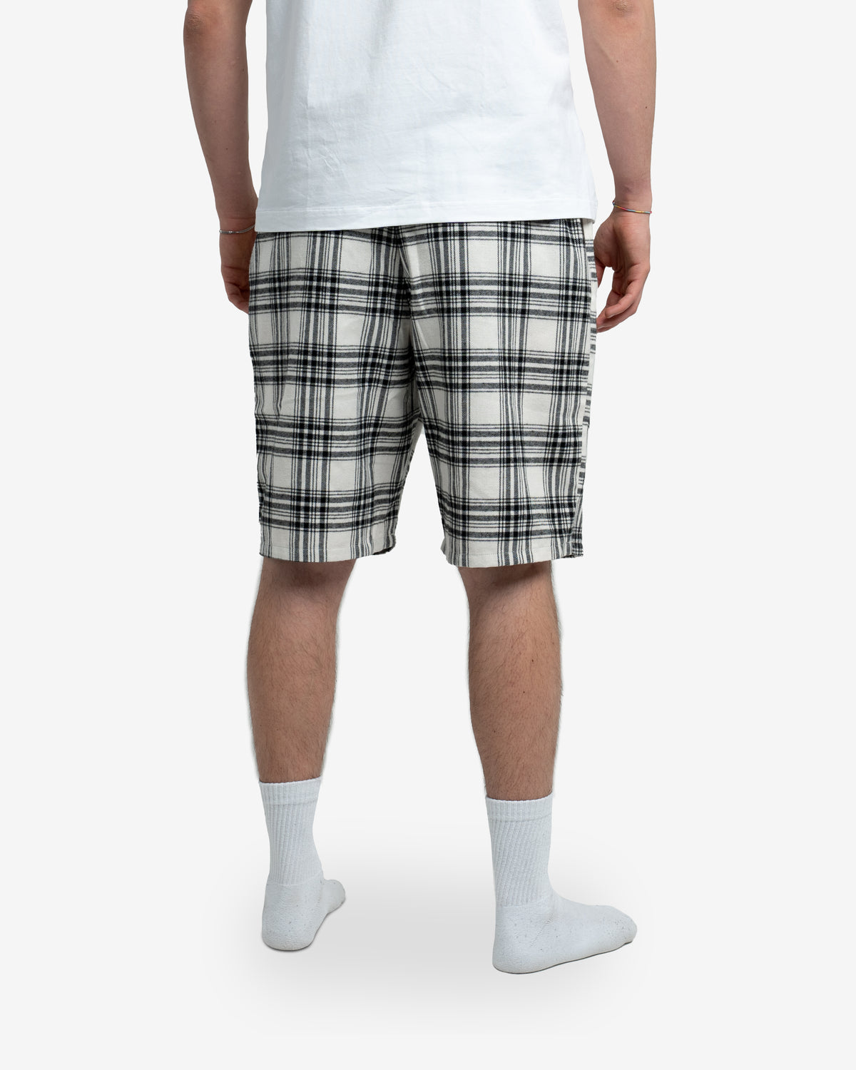 Modell steht mit dem Rücken zur Kamera und zeigt die Rückseite der Schorti in Creme mit schwarzem Karomuster. Weißes T-Shirt darüber, weiße Socken an den Füßen. Fokus auf Schnitt und Rückansicht der Shorts.