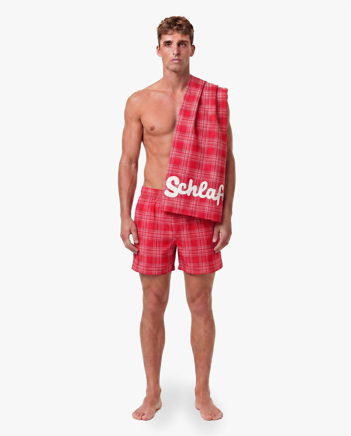 Model trägt rot karierte Shorts und präsentiert das passende Strandhandtuch mit weißem „Schlafi“-Schriftzug über der Schulter vor neutralem Hintergrund.
