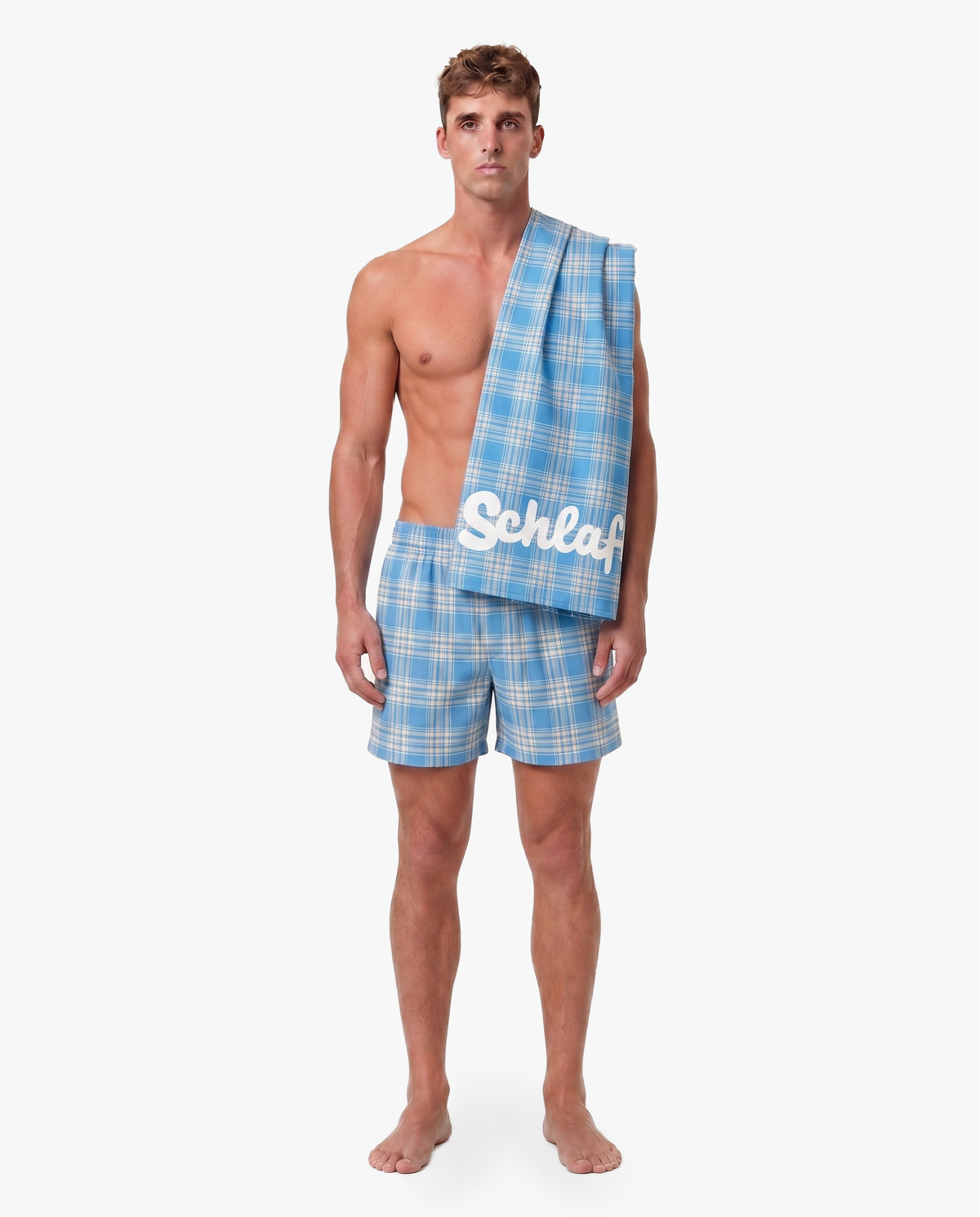 Model trägt blaue karierte Shorts und präsentiert das passende Strandhandtuch mit weißem „Schlafi“-Schriftzug über der Schulter vor neutralem Hintergrund.