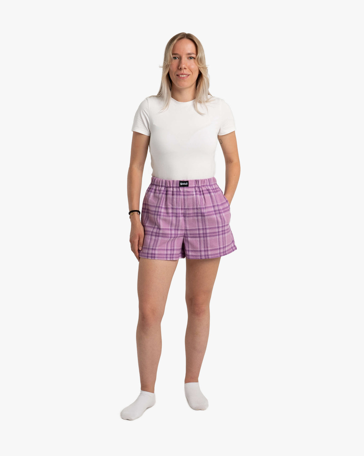 Eine Frau steht aufrecht vor weißem Hintergrund und trägt eine weiße, kurzärmlige T-Shirt und die SCHLAFI Hot Pants in der Farbe Wildberry – ein karierter Stoff in sattem Violett mit helleren und dunkleren Lilatönen. Die Shorts sitzen locker und enden kurz über dem Knie. Der elastische Bund sorgt für eine bequeme Passform, vorn ist ein schwarzes SCHLAFI-Label sichtbar. Die Frau trägt weiße Socken und lächelt dezent.