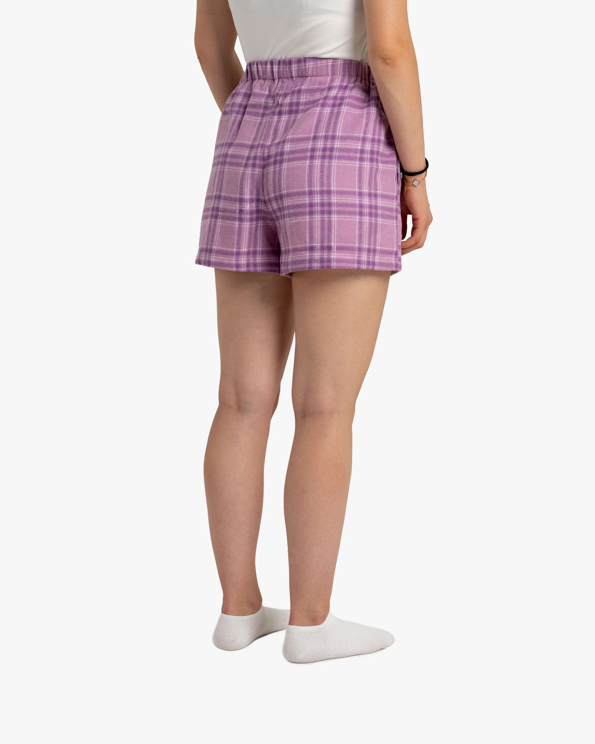 Rückansicht der Frau in derselben Hot Pants in Wildberry. Der karierte Stoff zieht sich gleichmäßig über die Rückseite, der Gummibund liegt gut an. Das Design zeigt keine Taschen oder sichtbaren Nähte, die Hose fällt locker über die Oberschenkel. Die Frau trägt weiße Sneakersocken.