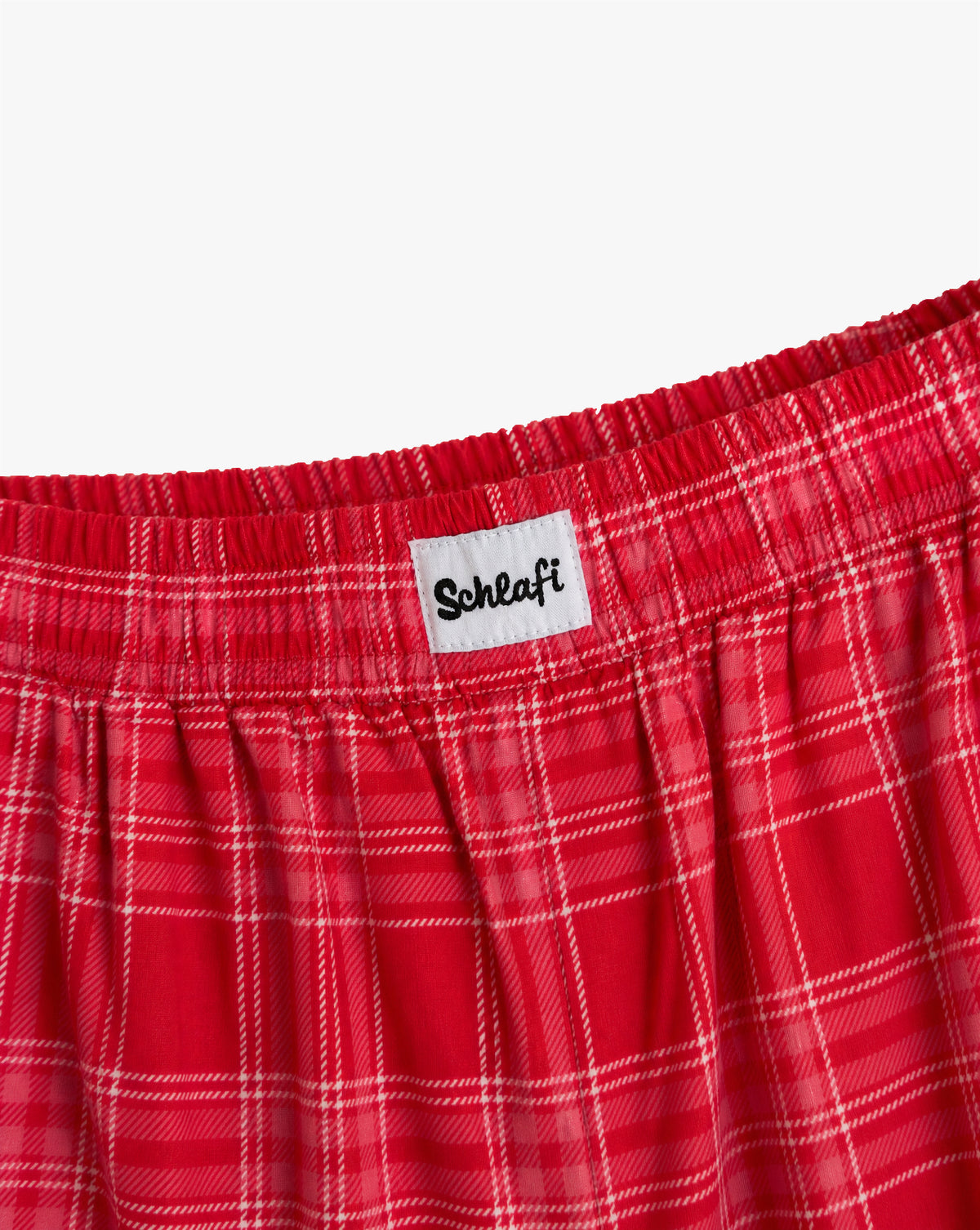 Alttext: Detailansicht des elastischen Bundes einer roten Badehose mit Karomuster. Weißes Label mit schwarzem „Schlafi“-Schriftzug auf der Vorderseite des Bundes.