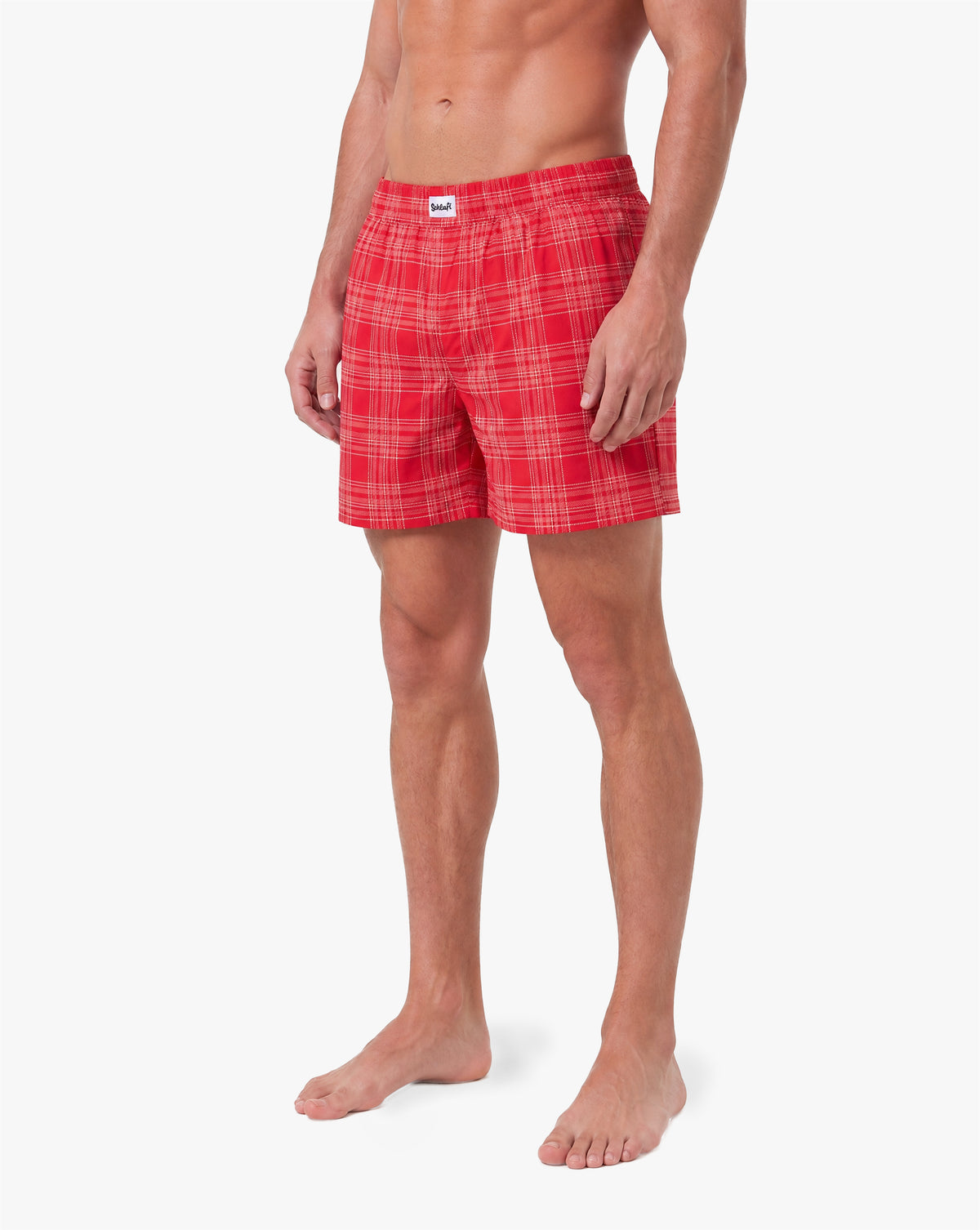 Alttext: Männliches Model trägt rote Badeshorts mit rosa-weißem Karomuster. Die Badehose verfügt über einen elastischen Bund und einen bequemen, lockeren Schnitt. Freigestellte Produktaufnahme vor grauem Hintergrund