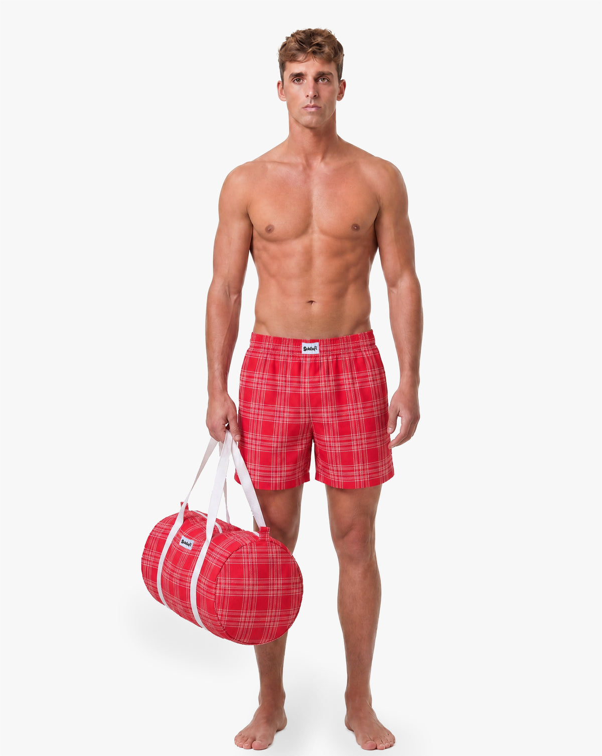 Model trägt rote karierte Schlafshorts und hält eine passende rote Reisetasche mit weißen Tragegriffen in der Hand.