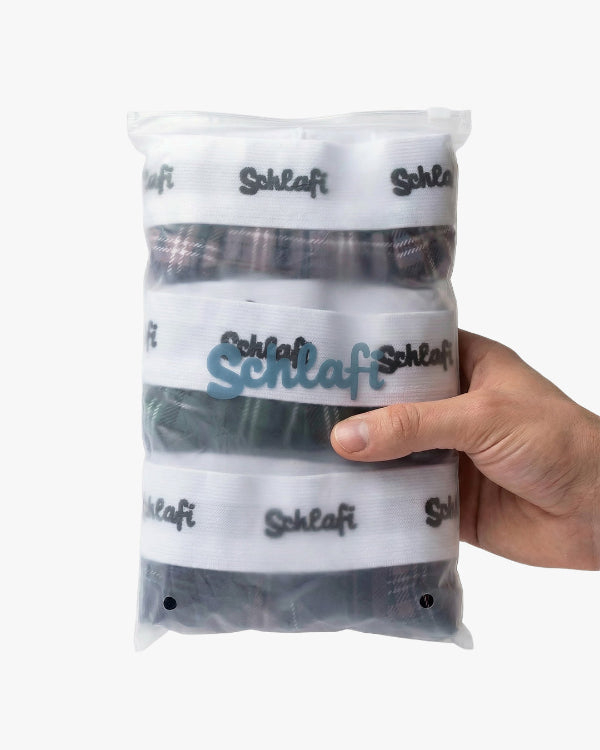 Dreier-Set „Schlafi“ Boxershorts in transparenter Reißverschluss-Tasche, von einer Hand gehalten. Sichtbar sind die karierten Designs und die weißen Logo-Bündchen.