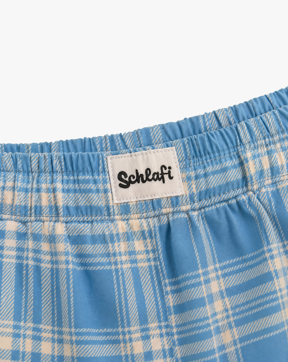Alttext: Detailansicht des elastischen Bundes einer hellblauen Badehose mit weißem Karomuster. Aufgenähtes weißes Label mit schwarzem „Schlafi“-Schriftzug auf der Vorderseite.