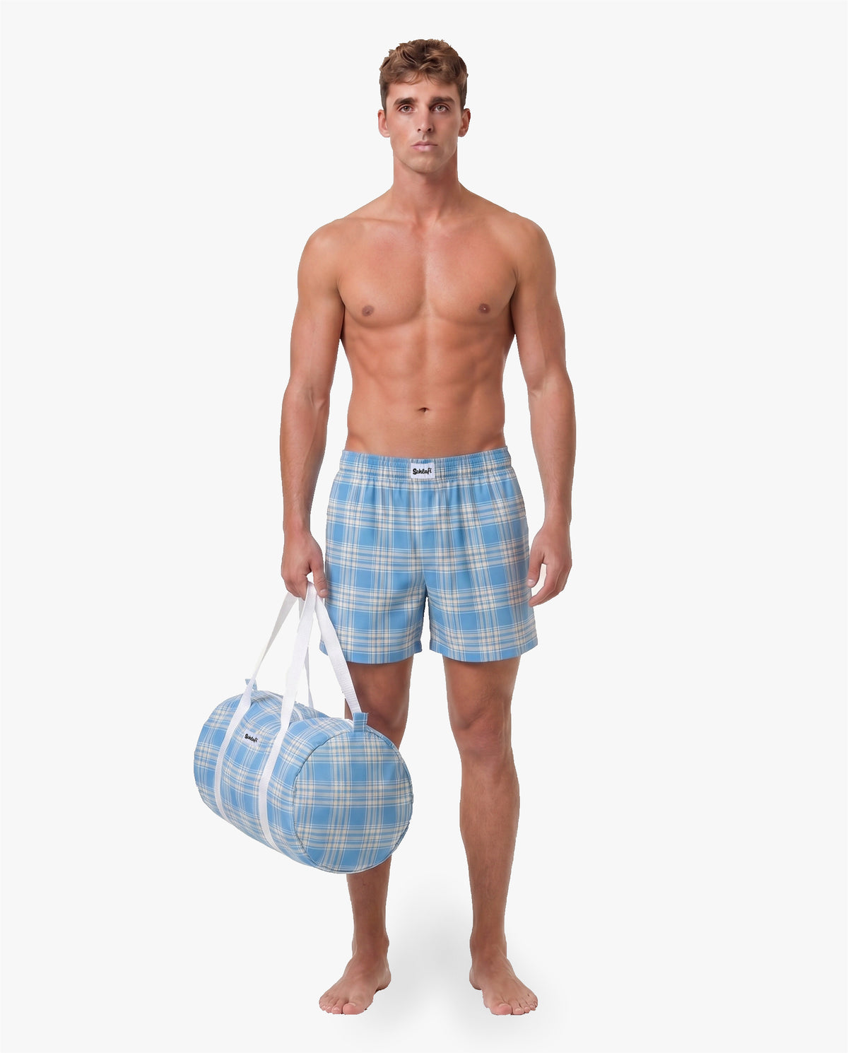 Model trägt hellblaue karierte Schlafshorts und hält eine passende hellblaue Reisetasche mit weißen Tragegriffen in der Hand.