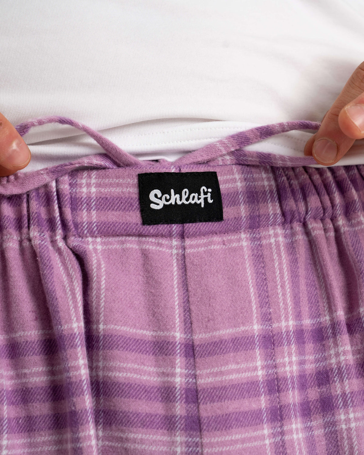 Nahaufnahme des Hosenbunds der SCHLAFI in Wildberry. Der Fokus liegt auf dem schwarzen SCHLAFI-Label und dem violetten Tunnelzug. Der Stoff zeigt das weiche Flanell-Karo in Lila- und Violetttönen. Die Hände halten den Bund locker auseinander.