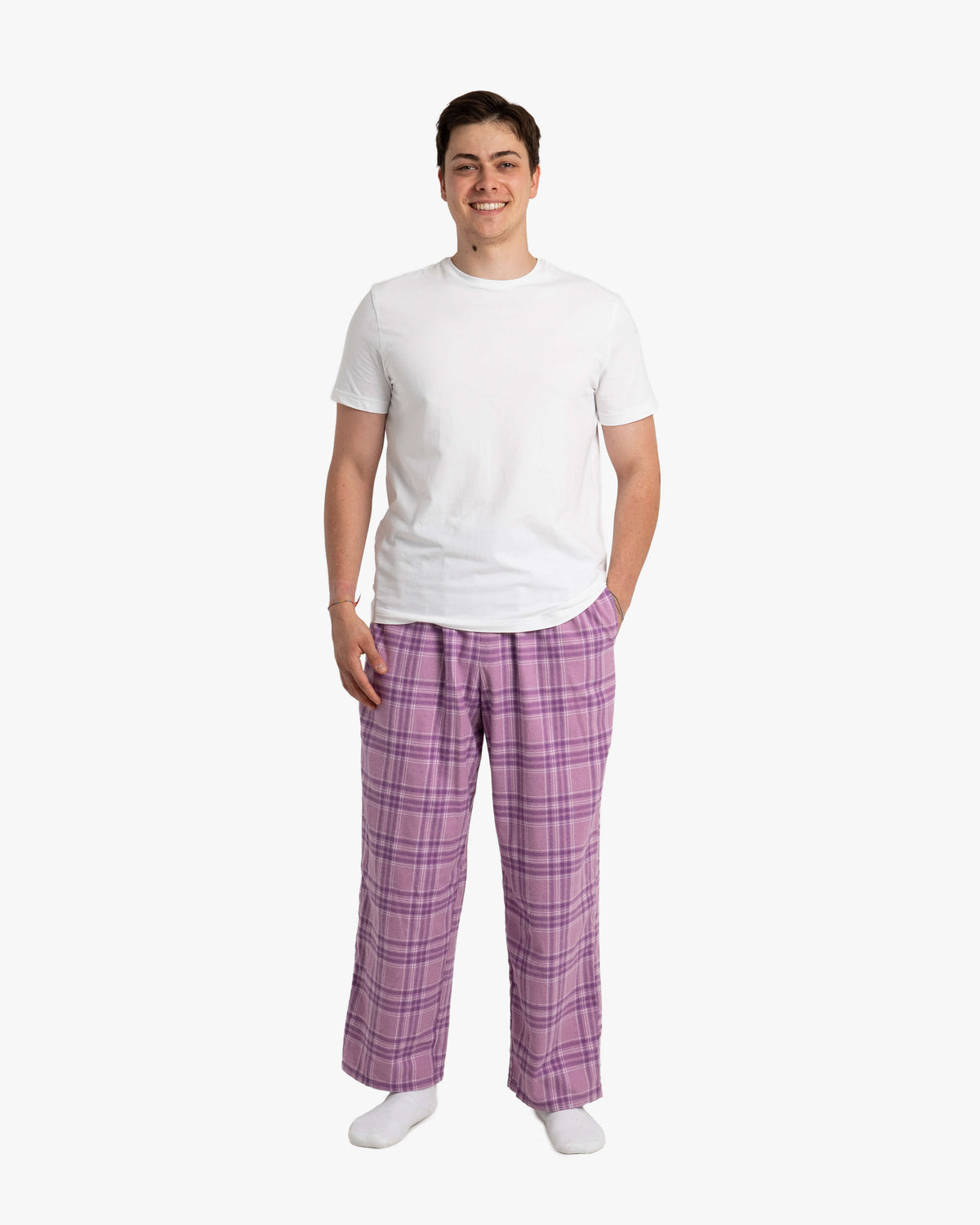 Mann steht vor weißem Hintergrund und trägt die lange SCHLAFI-Hose in Wildberry. Die Hose fällt locker und gerade bis zu den Knöcheln. Das Muster zeigt ein klassisches Karodesign in Violett- und Lilatönen. Dazu trägt er ein schlichtes weißes T-Shirt und weiße Socken. Er steht entspannt mit einem Lächeln, eine Hand locker in der Hosentasche.