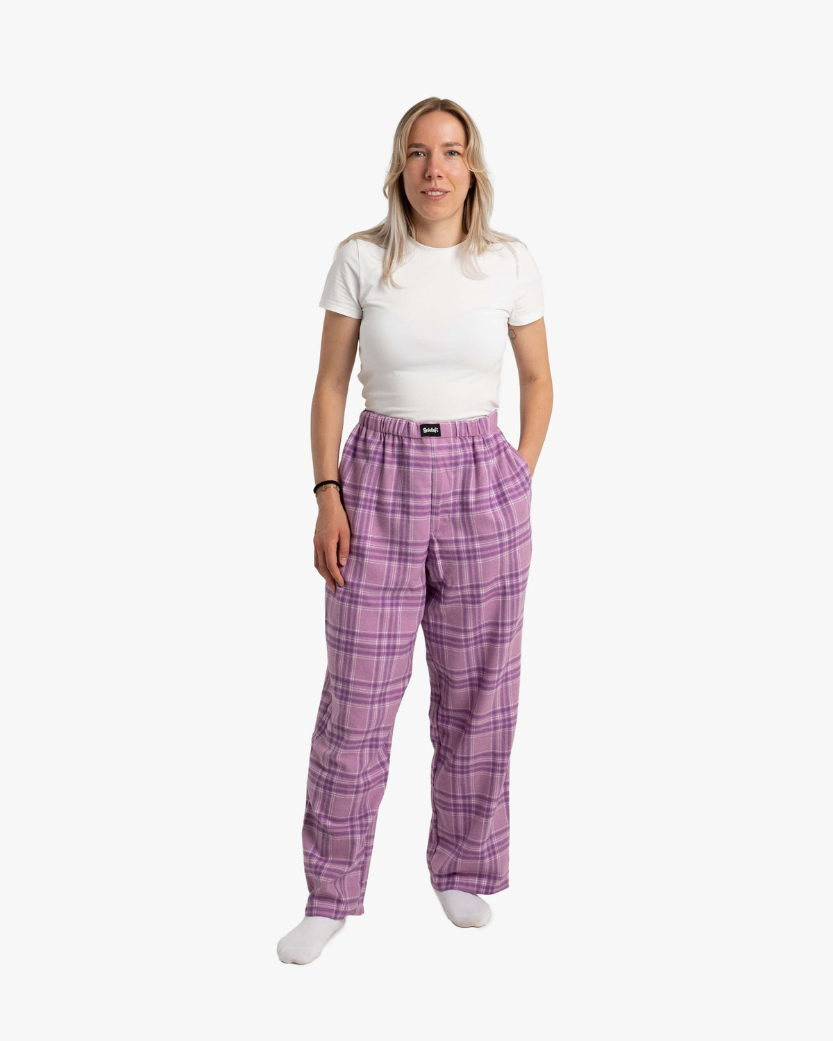Frau steht frontal zur Kamera und trägt die lange SCHLAFI-Hose in Wildberry. Das Karomuster aus Violett- und Lilatönen ist gleichmäßig verteilt. Die Hose sitzt hoch auf der Taille und fällt locker bis zu den Füßen. Sie kombiniert sie mit einem weißen, eng anliegenden T-Shirt und weißen Socken. Ihre Hände stecken lässig in den Hosentaschen.