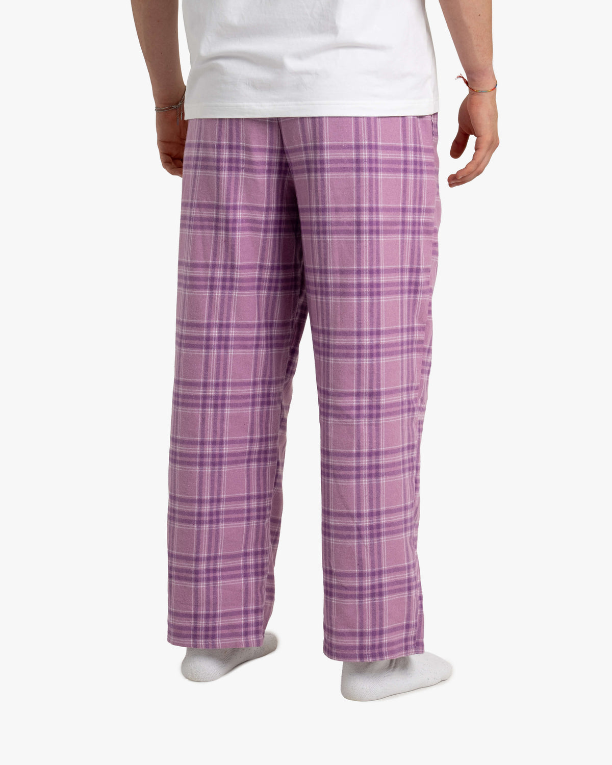 Mann steht mit dem Rücken zur Kamera. Die lange SCHLAFI-Hose in Wildberry fällt gerade über die Beine. Der elastische Bund liegt glatt an, das Karomuster verläuft gleichmäßig. Er trägt dazu weiße Socken und ein weißes Shirt, die Haltung ist entspannt.