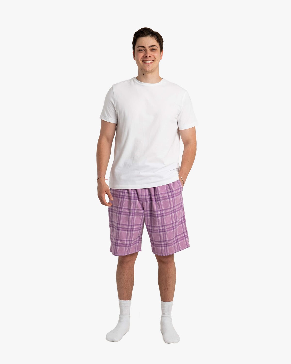 Mann steht frontal vor weißem Hintergrund und trägt die Schorti in Wildberry – knielange, locker fallende Shorts mit dezentem Karomuster in hellen und dunklen Beerentönen, kombiniert mit weißem T-Shirt und Socken.