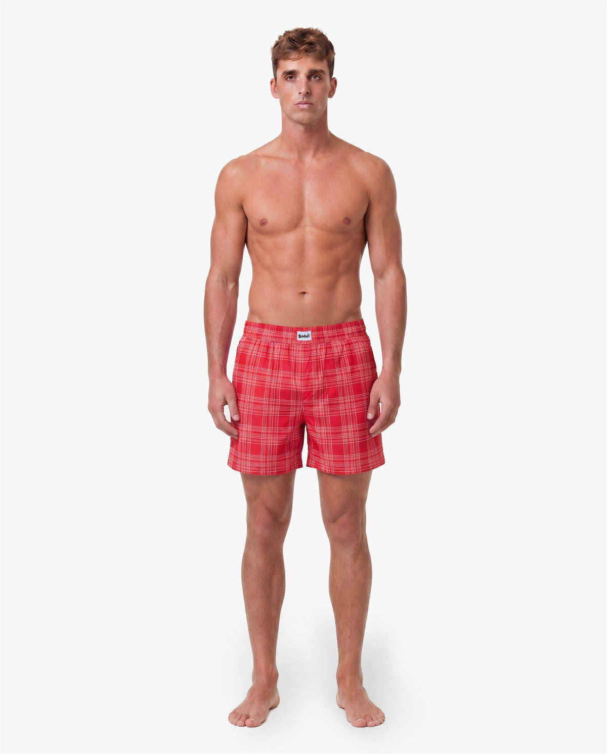 Vorderansicht eines männlichen Models in roten Badeshorts mit feinem Karomuster. Die Badehose besitzt einen elastischen Bund, seitliche Taschen und eine entspannte Passform. Freigestellte Produktaufnahme vor grauem Hintergrund