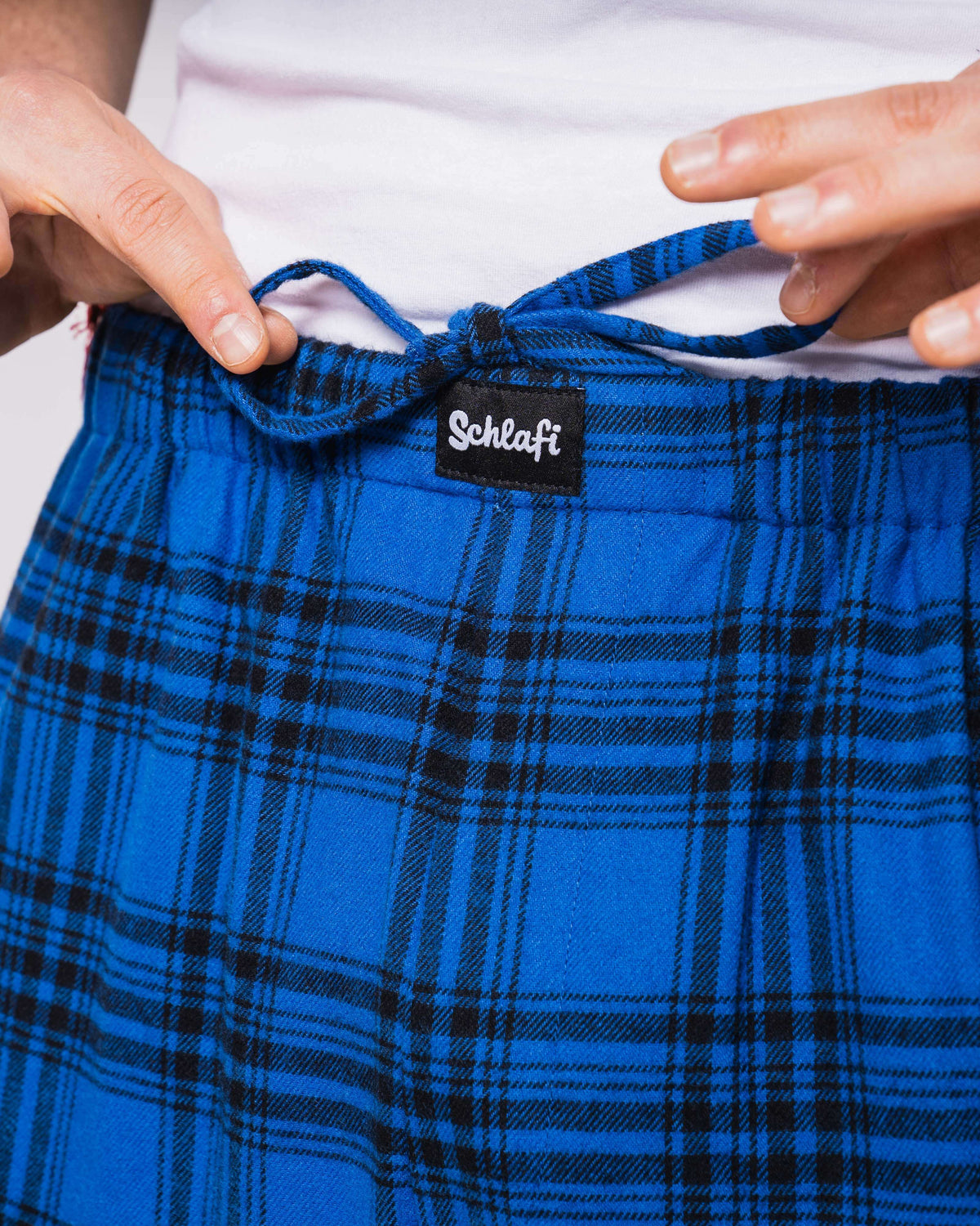 Nahaufnahme des Hosenbunds der Schlafi-Hose Blau mit sichtbarem „Schlafi“-Label und dunkelblauer Kordel. Die Hände des Models ziehen die Schleife leicht auseinander. Das Karomuster ist in kräftigem Königsblau mit schwarzen Linien gehalten.